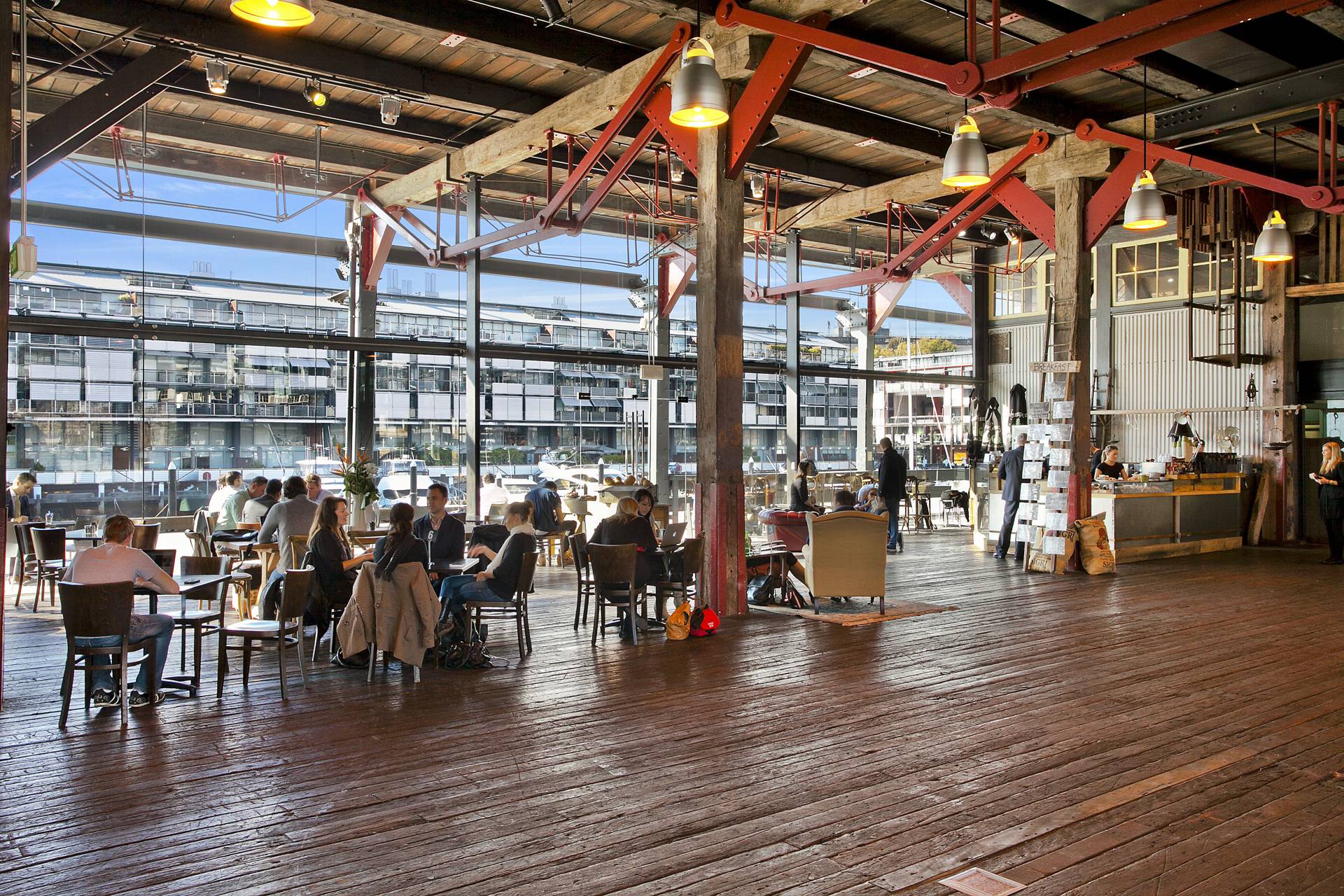 Diners at timber tables inside Elements Bar and Grill, under the wharf's exposed red trusses and ironbark columns, with the marina through full-height glazing